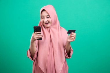 Heyecanlı tesettürlü Müslüman kadın cep telefonuyla online ödeme yapıyor ve elinde yeşil bir arka planda izole edilmiş bir kredi kartı tutuyor. Yeşil bir arka planda izole edilmiş, satın almasından memnun görünüyor..