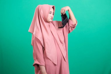 Sinirli bir kadın tesettür takmış iğrenç yüz maskesine bakıyor, yeşil bir arka planda izole edilmiş, muhtemelen pandemik, hijyenik olmayan ve tiksinmiş..