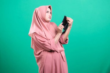 Başörtüsü olan Müslüman bir kadın, yeşil bir arka planda izole edilmiş mali durumu hakkında endişeli ve endişeli bir görünüm içinde boş cüzdanını inceliyor. Mali durumla başa çıkmanın zorluğunu gösteriyor. Bir borç sembolü..