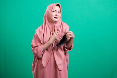 Yüzü maskeli, tesettürlü, tedirgin ve sağlıkla ilgili endişelerini dile getiren stresli bir Müslüman kadının portresi: yeşil bir arka planda izole edilmiş, zorluklar gösteren ve modern sağlık sorunlarıyla başa çıkan.