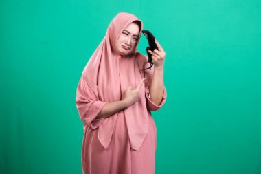 Başörtüsü takan, hayal kırıklığına uğramış ve gözlüklerine bakan, yeşil arka planda izole edilmiş rahatsızlık veya görme sorunları gösteren bir kadın..