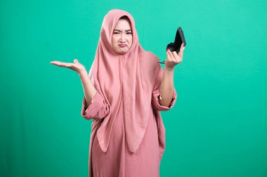 Başörtüsü takan hoşnutsuz bir kadın, yeşil bir arka planda izole edilmiş, ifadesel yüz ve el hareketleriyle kafa karışıklığı ve hayal kırıklığını ifade eden kırık bir akıllı telefon tutuyor..