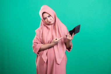 Başörtüsü takan bir kadın, yeşil bir arka planda izole edilmiş, mali mücadele ve durgunluğu sembolize eden hayal kırıklığına uğramış bir ifadeyle boş cüzdanını gösteriyor. Bu durum hayal kırıklığı ve belirsizliği çağrıştırıyor..