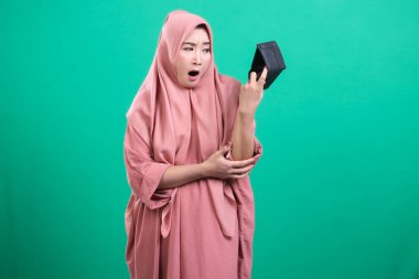 Başörtüsü olan endişeli müslüman kız yeşil bir arka planda soyutlanmış mali yıkım ve endişe ifadesiyle boş cüzdanına bakıyor..
