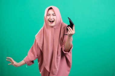 Genç Müslüman kadın, elinde boş bir cüzdan tutarken üzgün görünüyor. Stüdyo portresi ekonomik zorluklar veya maddi kayıplar, yeşil bir arka planda izole edilmiş..