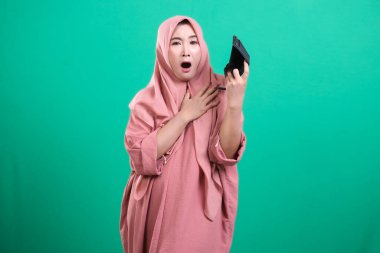 Boş bir cüzdan tutan, şok olmuş, tesettürlü Müslüman bir kız. Dehşet ve hayal kırıklığıyla tepki veriyor. Yeşil bir arka planda izole edilmiş, para kaybını resmediyor. Yeşil bir arkaplanda izole edilmiş.