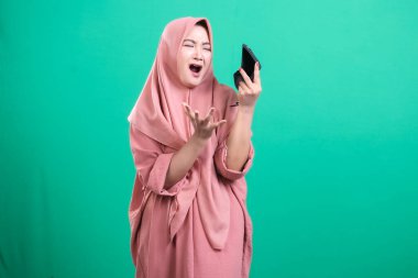 Cep telefonuyla hayal kırıklığını ifade eden İslamcı kadın, yeşil arka planda izole edilmiş haberlere olumsuz tepki gösteriyor. Modern teknolojinin zorluklarını gösteriyor..