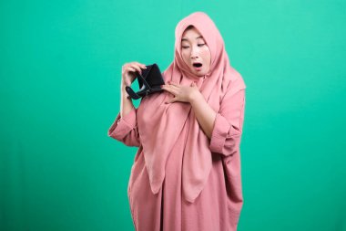 Başörtülü bir kadın, yeşil arka planda izole edilmiş mali zorlukları tasvir ederek boş cüzdanıyla şok ve hayal kırıklığını ifade eder. Bu resim potansiyel para sıkıntılarını ya da ani finansal zorlukları simgeliyor..