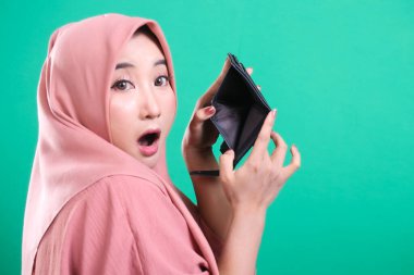 Sersemlemiş Müslüman kadın, yeşil arka planda izole edilmiş mali zorluk ve ekonomik zorlukları işaret ederek, boş cüzdanını tutarken şok olduğunu ifade ediyor..