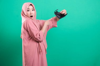 Yeşil bir arka planda izole edilmiş, maddi sıkıntılar nedeniyle boş cüzdanına bakan, şaşırmış bir ifadeyle genç Müslüman bir kadının portresi..