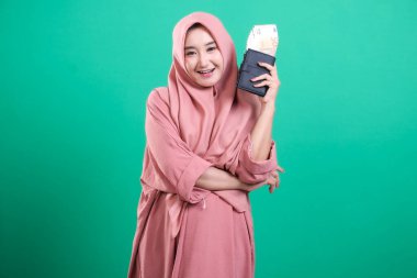 Başörtüsü takmış kendine güvenen Müslüman bir kadın yeşil bir arka planda izole edilmiş koyu bir cüzdanın içinde avro para birimini gösteriyor. Gülümsemesi iyimserlik ve finansal bağımsızlık yayıyor..