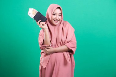 Avro banknotlarıyla dolu bir cüzdan taşıyan gülümseyen Müslüman bir kadın, yeşil bir arka planda izole edilmiş zenginlik ve refahı sembolize eden mutlu ve başarılı görünüyor. Finansal özgürlüğü ve ekonomik başarıyı ifade ediyor..