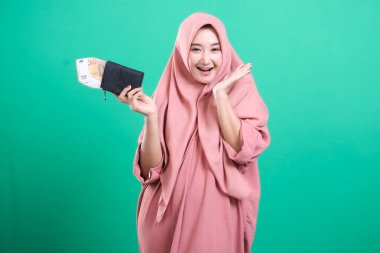 Geleneksel giyinmiş neşeli Müslüman bir kadın yeşil bir arka planda izole edilmiş mutluluk ve zenginliği ifade eden bir cüzdan dolusu para taşıyor. Mali durumu ve gelecekteki başarısı hakkında heyecanlı görünüyor..