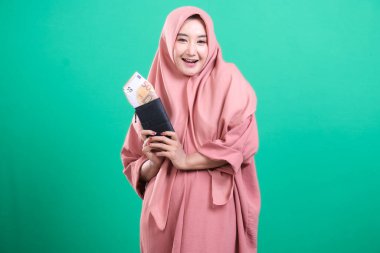 Tesettürlü neşeli Müslüman kadın para dolu bir cüzdan tutuyor ve gülümsüyor. Yeşil arka planda izole edilmiş finansal refah ve İslami bankacılık belirtileri.
