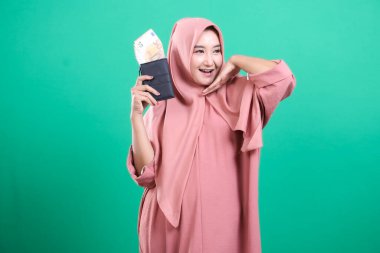 Neşeli Müslüman kadın para dolu bir cüzdanı elinde tutarak servetini sergiliyor ve gülümsüyor. Yeşil bir arka planda izole edilmiş finansal başarıyı simgeliyor. İslami finansal gücü somutlaştırıyor..