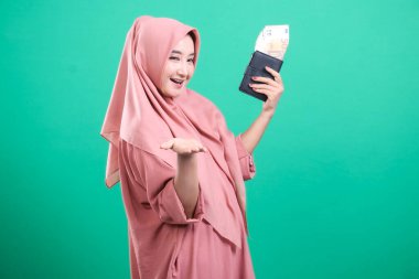 Mutlu Müslüman kadın tesettüre girmiş, elinde para dolu bir cüzdan, geniş bir gülümseme, heyecanlı görünüyor. Yeşil bir arka planda izole edilmiş. Bu görüntü kişisel finans, bolluk ve başarıyı temsil ediyor..