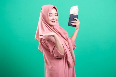 Yeşil bir arka planda izole edilmiş, tesettüre ve olumlu bir jestle, avro para birimi banknotlarıyla dolu bir cüzdan tutan neşeli Müslüman kadının portresi.