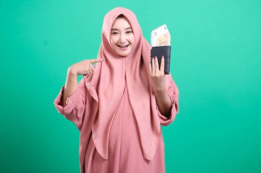 Para dolu cüzdan taşıyan, gülümseyen ve kendinden emin bir müslüman kızın portresi, yeşil bir arka planda izole edilmiş, finansal okuryazarlık becerileri gösteriyor. Yatırımlara ve finansal refaha karşı olumlu bir tutum sergiliyor.