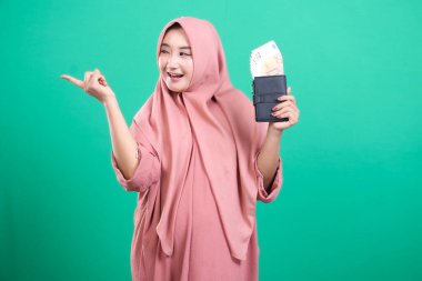 Güçlü Müslüman kadın, yeşil bir arka planda izole edilmiş, para dolu bir cüzdan ile para dolu, yatırım ve refahı sembolize ediyor..