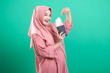 Başörtüsü olan mutlu genç bir kadın cüzdanını parayla dolduruyor. Yeşil bir arka planda izole edilmiş mali başarı ve mutluluğu simgeliyor..