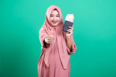 Pembe tesettürlü neşeli müslüman kadın, yeşil arka planda izole edilmiş, mali başarı ve mutluluğu sembolize eden, dolu bir cüzdan taşıyor. Modern İslami yaşam tarzında finansal bolluk.