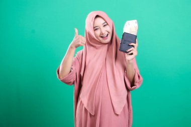 Başparmağıyla tesettürlü neşeli bir kadın, para dolu bir cüzdanla caka satıyor, yeşil bir arka planda izole edilmiş, yeni keşfedilen finansal başarı ve refahı ifade ediyor..