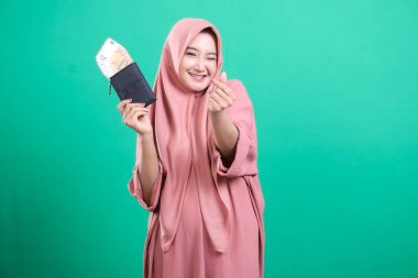 Başörtüsü takan genç Müslüman bir kadın yeşil bir arka planda izole edilmiş mutluluk ve başarıyı ifade eden para dolu bir cüzdanı tutarken geniş bir gülümseme içinde..