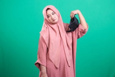 Hoşnutsuz bir kadın tesettüre bürünmüş, yüz maskesi takıyor, belirgin bir hoşnutsuzluk ve üzgün duygular taşıyor, yeşil bir arka planda izole edilmiş, korunmayla ilgili endişelerini dile getiriyor, 97 karakter.