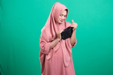 Boş cüzdanını gösteren tesettürlü genç bir kadın maddi zorluklar yaşıyor. Üzgün görünüyor. Ekonomik sorunları ve mali zorlukları sembolize etmek için uygun yeşil bir arka planda izole edilmiş.