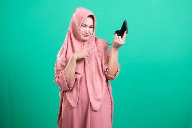 Tatmin olmamış yüzlü tesettürlü bir kadın ayakkabıyı inceliyor ve onu onaylamıyor, ürün kusuru gösteriyor, yeşil bir arka planda izole edilmiş. Görüntü onun olumsuz tepkisini ve dışlanmışlık ifadesini yakalıyor..