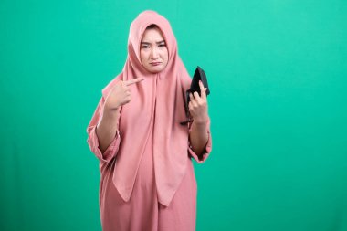 Üzücü ve stresli tesettürlü Müslüman kadın yeşil arka planda izole edilmiş, üzgün bir yüz ifadesi olan boş bir cüzdana işaret ediyor..