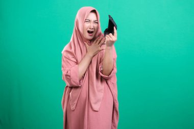 Genç Müslüman bir kadın, elinde boş bir cüzdan tutarken şok olmuş görünüyor. Yeşil bir arka planda, mali kriz konusunda çaresiz ve umutsuz hissediyor..