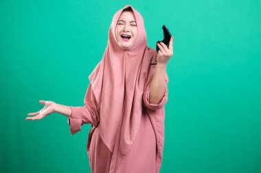 Sinyal yetmezliğinden dolayı kapalı bir telefon çağrısına bağıran tesettürlü, tesettürlü genç müslüman bir kadın, yeşil bir arka planda izole edilmiş. Hayal kırıklığını ve güvensizliğini el hareketleriyle ifade ediyor..