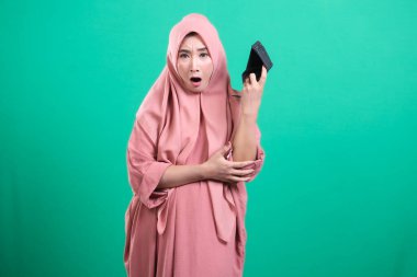 Boş bir cüzdan sergileyen umutsuz Müslüman bir kadın. Yeşil arka planda izole edilmiş, maddi sıkıntıyı simgeliyor. İnsanların karşılaştığı finansal zorlukları tasvir ediyor. Üzgün ve endişeli..