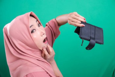 Yeşil arka planda soyutlanmış, boş cüzdanını gösteren tesettürlü bir kadın endişeli görünüyor. Finansal kaygıları gözler önüne seren imaj, ekonomik istikrarsızlık ve endişeye yol açıyor.