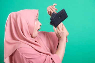 Şok olmuş, tesettürlü Müslüman bir kadın yeşil arka planda soyutlanmış, maddi sıkıntı ve umutsuzluğu ifade eden boş cüzdanına bakar. Üzücü bir ifadeyle para sorunlarıyla karşı karşıya ve geliri yok..
