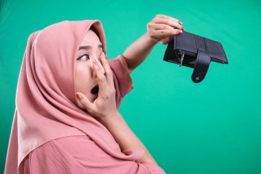 Başörtüsü takan bir kadın, yeşil arka planda izole edilmiş, korkunç bir ifadeyle boş cüzdanına bakar. Bu durum yoksulluk ve ekonomik güvensizlikle ilgili kaygıları vurguluyor.