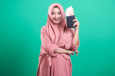 Avro banknotlarıyla dolu cüzdanı olan tesettürlü mutlu Müslüman kadın ve yeşil arka planda izole edilmiş kameraya gülümsüyor. Tasarruflarıyla refah, başarı ve mali güvenliği gösteriyor..