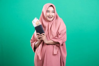 Avro banknotlarıyla dolu bir cüzdanı olan tesettürlü Müslüman bir kadın, yeşil arka planda, en az 90 karakter, en fazla 110 karakter ile izole edilmiş mali geleceği hakkında iyimser görünüyor.