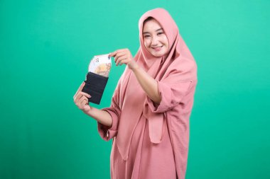 Pembe başörtüsü içinde gülümseyen genç muslimah avro banknotları ile bir cüzdan gösteriyor, yeşil bir arka planda izole mali refah ve ekonomik istikrar tasvir ediyor.