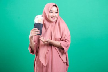 Başörtüsü takan mutlu genç bir kadın, yeşil arka planda izole edilmiş avro banknotlarıyla dolu bir cüzdanı tutarak servetini sergiliyor. Gururlu ve başarılı görünüyor..