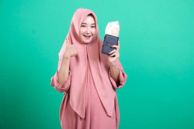 Tesettürlü neşeli Müslüman kadın yeşil bir arka planda izole edilmiş mali başarıyı sembolize eden para dolu bir cüzdanla başarı gösteriyor. Bu resim olumlu finansal beklentiler ve refahı gösteriyor.