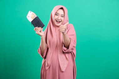 Zevkli tesettürlü kadın cüzdanını parayla şişirerek yeşil arka planda izole edilmiş başarı işaretini veriyor. Mali bağımsızlık ve yatırım başarısı kavramı.