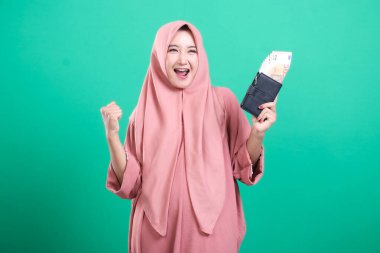 Para dolu cüzdanı olan neşeli, genç Müslüman bir kadın yeşil bir arka planda izole edilmiş, mutlu bir gülümsemeyle finansal başarıyı kutluyor. Parasal kazancı ya da zaferi onu mutlu ediyor.