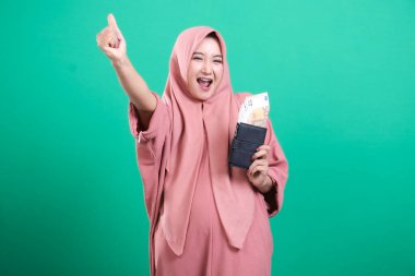 Avro para dolu bir cüzdan taşıyan tesettürlü Müslüman bir kadın yeşil arka planda izole edilmiş bir şekilde onay veriyor. Finansal başarısını kutluyor. Çok mutlu..