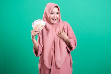 Başörtüsü takan genç bir kadın, elinde heyecanlı bir ifadeyle, yeşil bir arka planda izole edilmiş, finansal başarı gösteren bir para tutuyor. Zenginliğiyle mutlu görünüyor..