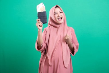 Başörtülü mutlu bir Müslüman kadın, cüzdanını avro dolu tutarak mali refahını kutluyor. Servetinden çok memnun, yeşil bir arka planda izole edilmiş..