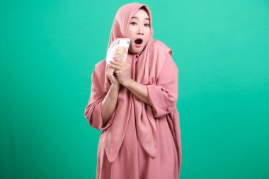 Tesettürlü genç kadın, elinde avro banknotları tutarken hoş bir şaşkınlık yaşıyor. Yeşil arka planda izole edilmiş bir mali servet ve neşe portresi. Yüz altı karakter kullanıyor.