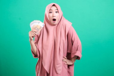 Heyecanlı tesettürlü Müslüman bir kadın elinde bir yığın avro banknotuyla, yeşil bir arka planda izole bir şekilde mali başarı hissini iletiyor. Şaşırdığını ve sevindiğini ifade ediyor. Beklenmedik bir zenginlik öneriyor..