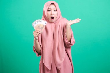 Elinde avro banknotları tutan tesettürlü neşeli Müslüman bir kadın yeşil bir arka planda izole edilmiş keyifli bir şok ve şaşkınlık ifadesi sergiliyor. Elindeki döviz miktarına şaşırdı..